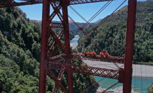 A group of five construction workers in orange safety gear are sitting on the metal beams of a large red bridge high above a scenic river valley. The workers discuss against lush green hills under a clear blue sky.