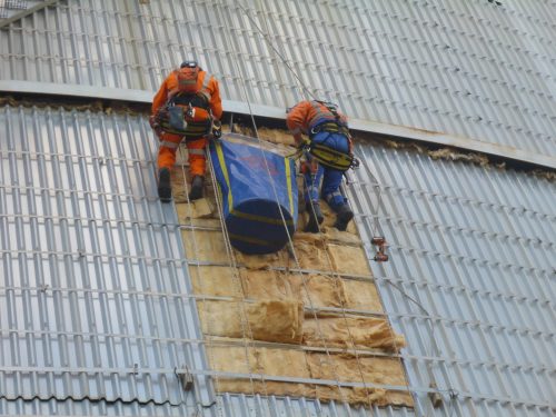 Two rope access technicians in high-visibility orange and blue gear are performing repairs on the exterior of a large corrugated metal building. They are secured with harnesses and ropes, carefully working side by side as they install insulation material under the building's cladding. The photo captures them in action, demonstrating their work's skilled and precarious nature at considerable height.