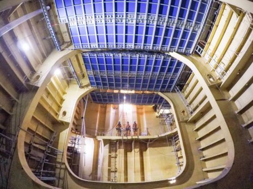 Interior view of a large ship under construction, showcasing a spacious and intricately designed compartment. The ceiling features a grid of blue panels, while scaffolding and ladders are positioned along the walls, indicating ongoing work. In the distance, three workers can be seen standing on a raised platform, illuminated by the overhead lights, providing a sense of scale and highlighting the complexity of the shipbuilding process.