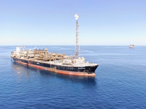 A serene aerial view of the FPSO vessel 'Raroa' positioned in the calm blue ocean under a clear sky. The vessel, painted in black with a red lower hull, features an extensive array of industrial equipment and machinery on its deck, including a tall flare stack actively burning off gas. In the distance, another similar vessel can be seen, highlighting the scale of offshore operations in this region.