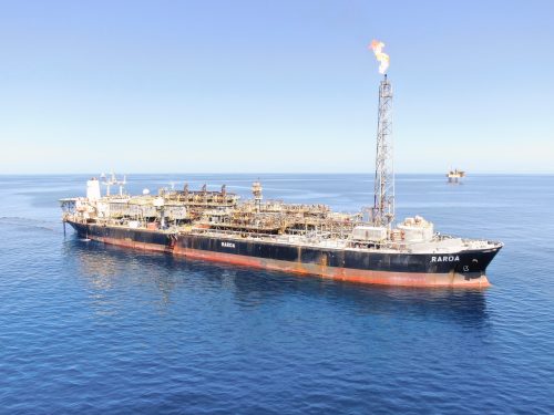 A serene aerial view of the FPSO vessel 'Raroa' positioned in the calm blue ocean under a clear sky. The vessel, painted in black with a red lower hull, features an extensive array of industrial equipment and machinery on its deck, including a tall flare stack actively burning off gas. In the distance, another similar vessel can be seen, highlighting the scale of offshore operations in this region.