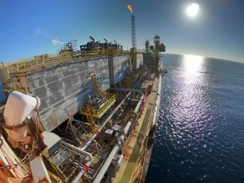 Spectacular wide-angle view of an FPSO. The vessel, heavily equipped with industrial machinery and piping, shows signs of weathering and rust. A flare stack actively burns in the background, glowing against the setting sun—the sea sparkles beneath the sunlight, highlighting this energy production facility's vast and isolated environment.