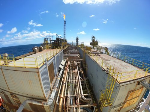 A wide-angle view from an offshore oil platform's deck shows a complex network of pipelines, safety railings, and operational machinery under a clear blue sky. A flare stack is actively burning off the gas at the left, highlighting the ongoing production process. The ocean extends into the horizon, reflecting the sunlight and emphasising the isolated location of this energy-producing facility.