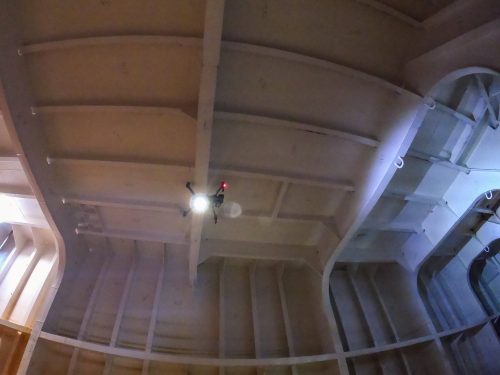 Interior view of a ship's curved ceiling structure, showcasing robust, arched steel beams painted in light grey. In the centre, a drone hovers, emitting bright light from its two glowing bulbs and a red indicator light, illuminating the surrounding area. The image captures the engineering detail and the vastness inside the ship, highlighting the innovative use of drones in the construction process.