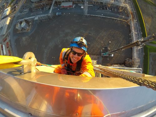 A close-up perspective of a smiling rope access technician in an orange suit and blue helmet, securely attached with safety gear atop an extremely high industrial structure. The camera angle captures the technician above, showcasing a vertiginous view of the ground far below, where industrial equipment and vehicles are minuscule. The setting sun casts a warm glow, emphasising the height and risky nature of the work environment.
