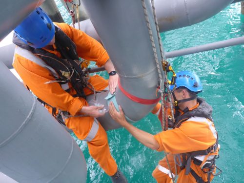 Two Vertech Group technicians in orange high-visibility suits and safety harnesses perform maintenance work on a large metallic structure suspended above turquoise waters. The technician in the foreground wears a blue helmet, while the other wears a light blue helmet. Both are securely attached to the structure with safety ropes as they apply protective tape around a joint, demonstrating meticulous attention to detail in their specialist access work.