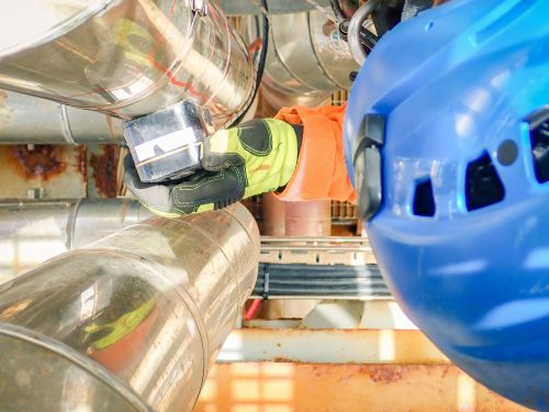 Close-up view of a Vertech Group technician conducting ultrasonic testing on a metallic pipeline. The technician, wearing a blue helmet and orange safety gloves, holds a small ultrasonic device against the pipe. The device's screen displays data crucial for assessing the pipeline's integrity. This detailed inspection ensures the pipeline's safe and efficient operation within the industrial facility.