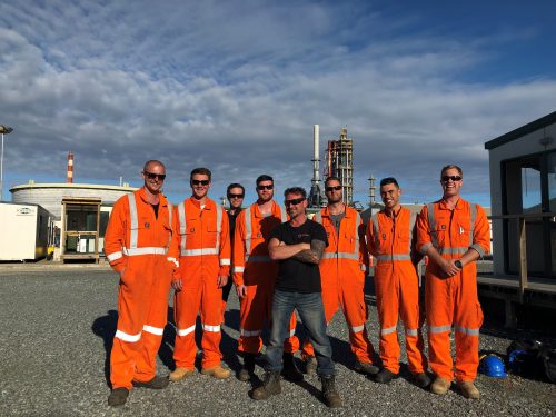 A group of eight Vertech Group employees stands together in front of an industrial facility, posing for a team photo under a clear sky. Seven team members are dressed in bright orange high-visibility coveralls, while one is wearing a black Vertech T-shirt. They all display a sense of camaraderie, smiling and relaxed, highlighting the unity and professionalism of the team.