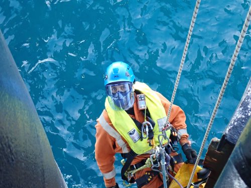 Close-up of a technician wearing an orange suit and a blue helmet, outfitted with a full-face visor and a bright yellow life vest, as he works on an offshore rig. He is secured with multiple safety ropes, hanging above the dark, rippling ocean waters. His gear includes various tools and safety devices attached to his harness, ready for use in this critical and hazardous maintenance operation.