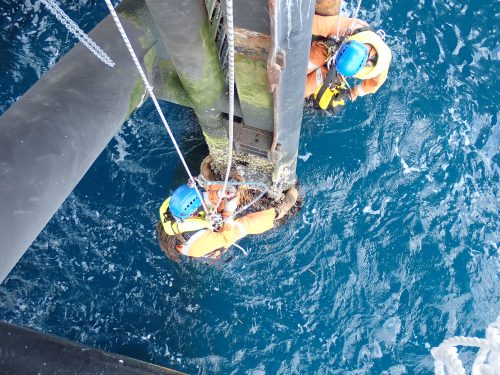 Ropes suspend two technicians in safety gear as they work closely together on the structural components of an offshore oil platform. Positioned directly over the deep blue sea, they focus on equipment attached to one of the platform's large vertical pillars. Their bright orange suits and blue helmets stand out against the vivid colour of the ocean, illustrating the demanding and precise nature of their tasks in such a challenging environment.