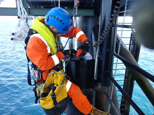 A focused technician in an orange suit and blue helmet, equipped with a safety harness, is performing maintenance on the structural elements of an offshore oil rig. Suspended above the ocean, he uses tools from his yellow utility bag to work on a large black column, part of the rig's support structure. The backdrop features the ocean's deep blue waters, enhancing the isolation and critical nature of the work environment.