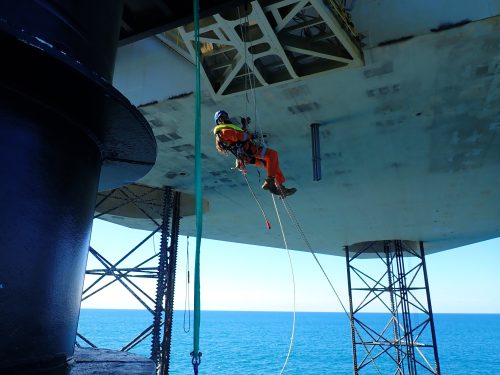 A technician in an orange suit and yellow helmet is suspended by ropes beneath an offshore platform, performing maintenance work. He is surrounded by the vast blue sea, with the platform's massive grey structure overhead. The technician uses various tools as he works on the underside of the platform, demonstrating the critical and high-skilled nature of rope access tasks in such challenging environments.