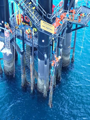 An overhead view of technicians working on an offshore oil rig platform above the clear turquoise waters. The workers, dressed in orange safety gear, perform maintenance tasks on the rig's structural pillars. The scene includes various safety signs, bright yellow guardrails, and an array of equipment, highlighting the complexity and stringent safety protocols necessary for such operations.
