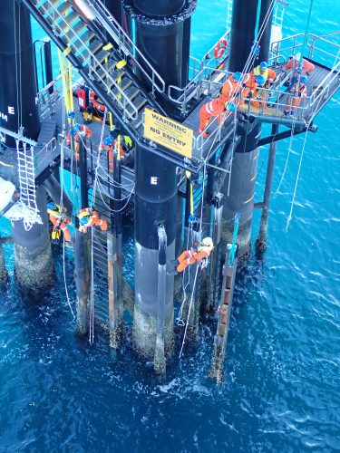 An overhead view of technicians working on an offshore oil rig platform above the clear turquoise waters. The workers, dressed in orange safety gear, perform maintenance tasks on the rig's structural pillars. The scene includes various safety signs, bright yellow guardrails, and an array of equipment, highlighting the complexity and stringent safety protocols necessary for such operations.