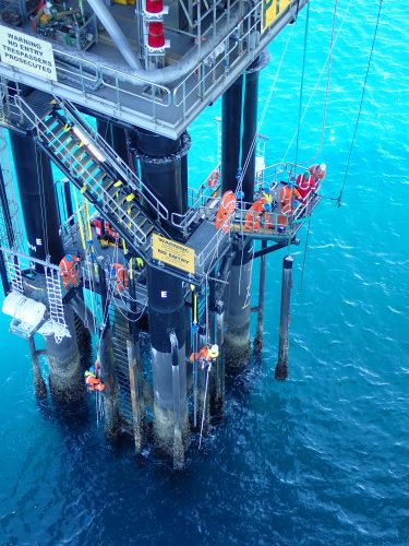 An overhead view of technicians working on an offshore oil rig platform above the clear turquoise waters. The workers, dressed in orange safety gear, perform maintenance tasks on the rig's structural pillars. The scene includes various safety signs, bright yellow guardrails, and an array of equipment, highlighting the complexity and stringent safety protocols necessary for such operations.