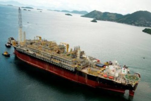 Aerial view of a large red and black floating production storage and offloading (FPSO) vessel stationed in a calm sea surrounded by green islands. The FPSO has extensive industrial facilities, including cranes, tanks, and various complex machinery on its deck, illustrating its capability for offshore oil and gas processing and storage.