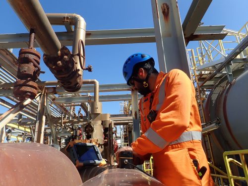 A Vertech Group technician in an orange high-visibility suit and blue helmet is intently using a handheld inspection device near an industrial facility's complex arrangement of rusted valves and pipelines. The portable monitor on the left shows real-time data, aiding the technician in the non-destructive testing process. This critical inspection occurs amidst an elaborate network of metal pipes and industrial structures under a clear sky.