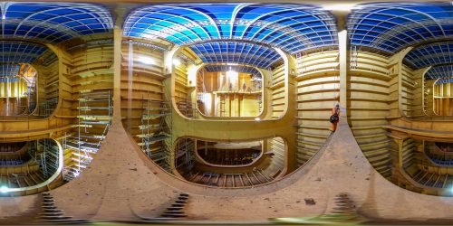 A panoramic and surreal view inside a ship's hull under construction, captured using a fisheye lens. The image features a kaleidoscopic effect with multiple mirrored and curved photos of the ship's interior. Steel beams painted blue and yellow create an intricate geometric pattern across the ceiling, while scaffolding and construction equipment are visible at various levels. A single technician in an orange suit is prominently positioned in the frame, accentuating the scale and complexity of the shipbuilding process.