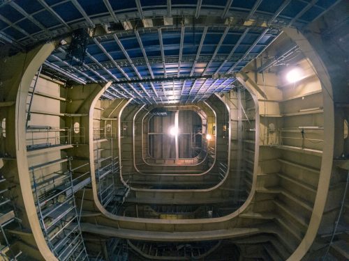 Looking upwards inside the cavernous hull of a ship, the photo captures the geometric beauty of multiple tiers of steel beams that form the ceiling. The beams are painted in alternating shades of blue and grey, creating a striking visual pattern. Bright work lights illuminate the structure, and a few technicians can be seen working on the distant platforms, emphasising the scale and complexity of the shipbuilding process.