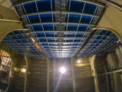 Looking upwards inside the cavernous hull of a ship, the photo captures the geometric beauty of multiple tiers of steel beams that form the ceiling. The beams are painted in alternating shades of blue and grey, creating a striking visual pattern. Bright work lights illuminate the structure, and a few technicians can be seen working on the distant platforms, emphasising the scale and complexity of the shipbuilding process.