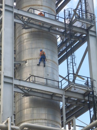 A rope access technician in orange and blue safety gear is performing maintenance work on the exterior of a large metal silo at an industrial facility. He is secured with safety harnesses and ropes as he descends the side of the silo, surrounded by an intricate framework of metal staircases and piping. Above him, another technician works from the metal platform, illustrating the precision and safety involved in high-altitude industrial operations.