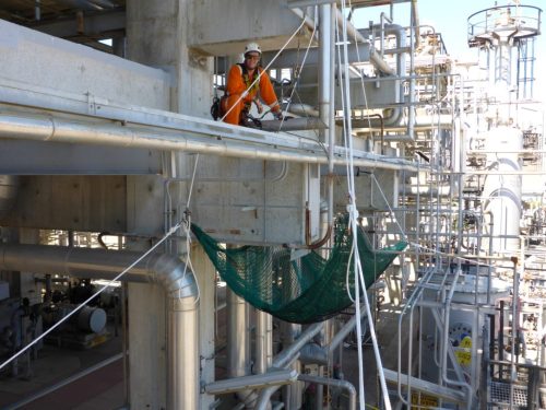 A technician in an orange high-visibility suit and white helmet is secured with safety harnesses while working on a rope access system within an industrial plant. He is positioned near a complex network of pipes and machinery, using a safety net for tool capture. The background is densely packed with various metal structures and operational equipment, showcasing the challenging environment for maintenance and inspection tasks.