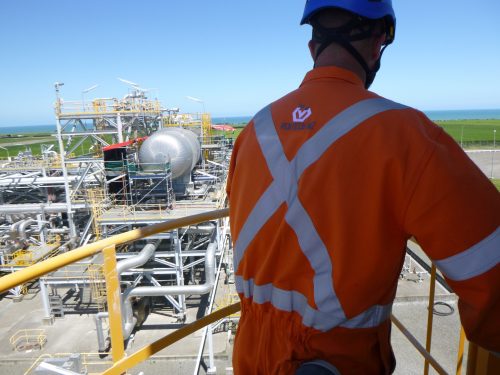 A Vertech Group employee stands on a Beach Energy Kupe Plant platform, overlooking the extensive industrial setup. He is dressed in an orange high-visibility jacket with reflective stripes and a blue safety helmet. The background features the coastal landscape near the plant, with a complex network of yellow pipes and large metallic structures visible below.